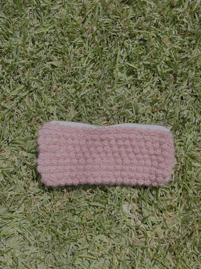 A pink bobble-stitch crochet pencil case lying closed on grass
